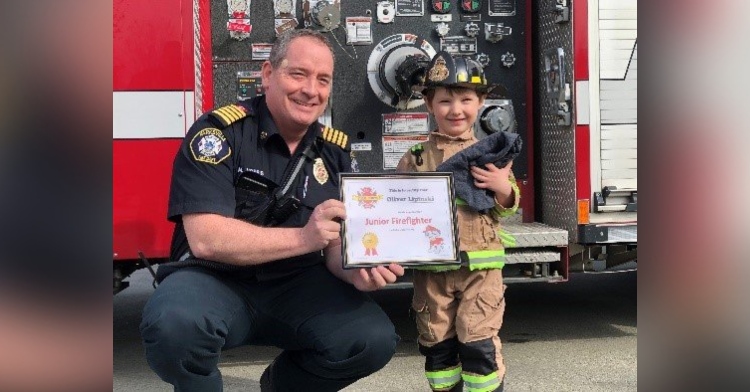 a firefighter from the parksville fire department smiling as he squats down next to 3-year-old oliver lipinski to present him with a certificate recognizing his status as an official junior member of the department. oliver is also smiling and is wearing full turnout gear.