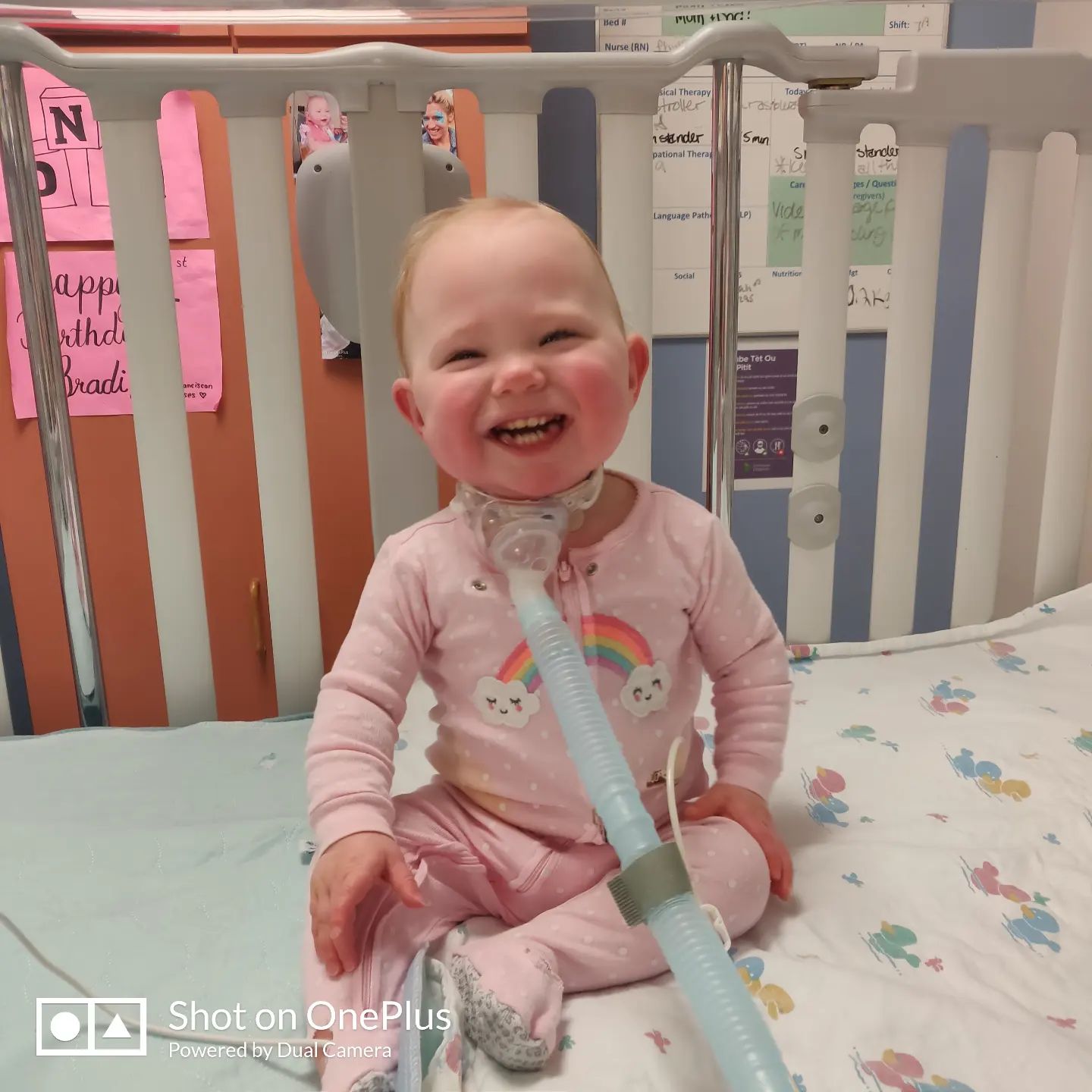 bradi foster smiling as she sits in a crib. she's wearing a gastrostomy tube.