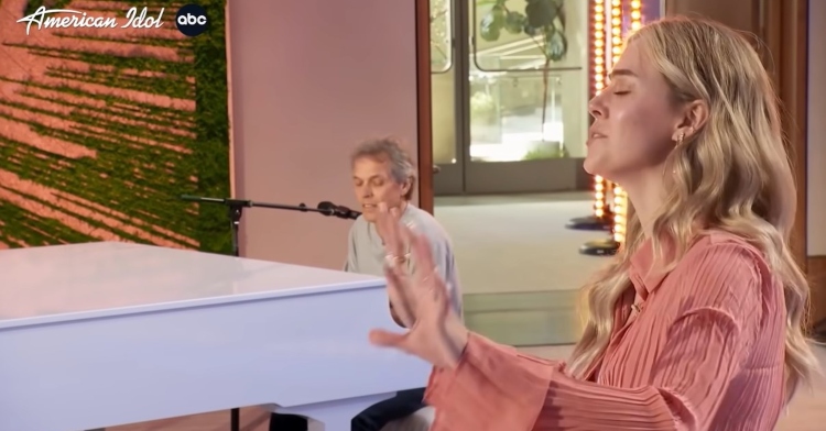 carly mickeal auditioning for “american idol.” she has her eyes closed and her hands extended in front of her. at a distance, her dad, dave, sits at a white piano to accompany her.