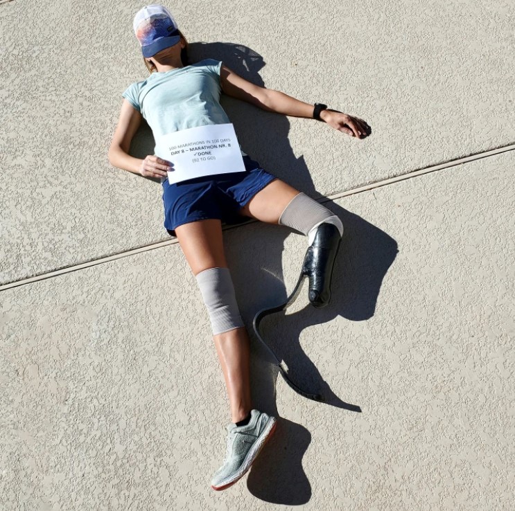 jacky hunt-broersma laying on the ground after completing a marathon. she has a baseball cap covering her face and she's holding a sign on her stomach. the sign reads 