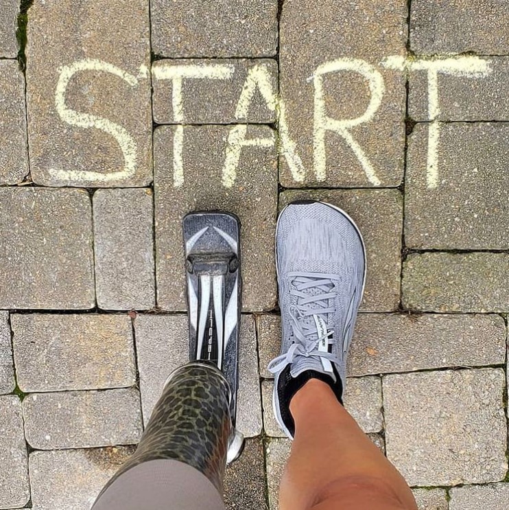 top-down view of a brick walkway that has the word "start" written on it in yellow chalk. just below the words are the feet of jacky hunt-broersma. on the right she is wearing a gray tennis shoe and on the left she is wearing her running prosthetic. 