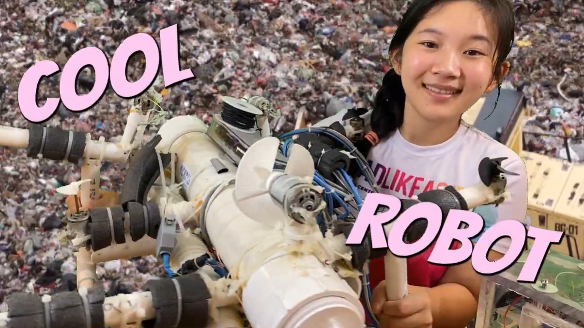 screenshot of a youtube video by anna du. in it she is smiling while holding her remote-operated vehicle (rov)  made to detect plastics on the ocean floor. the backdrop is of a large pile of garbage. the words "cool robot" have been edited onto the image.