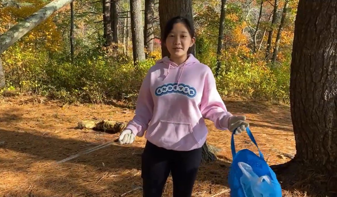 screenshot of a youtube video by anna du. she is standing in the woods, talking about picking up trash as she does just that. in one hand is a reusable bag full of trash and in the other is an object used to pick up the trash.