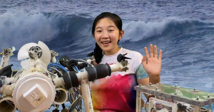 screenshot of a youtube video by anna du. she is smiling and waving. in front of her is her robot made to detect plastic on the floor of the ocean. the backdrop is of the ocean with the use of a green screen.