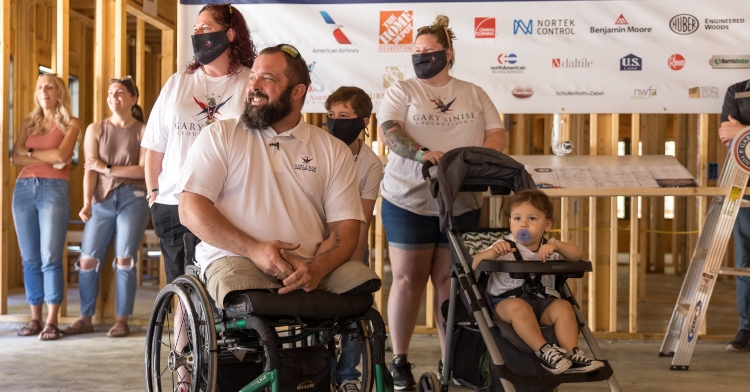 retired u.s. army sergeant christopher kurtz smiling with his wife heather and their child, along with other family and friends, as they stand inside the foundation of the home that’s being built for them by the gary sinise foundation