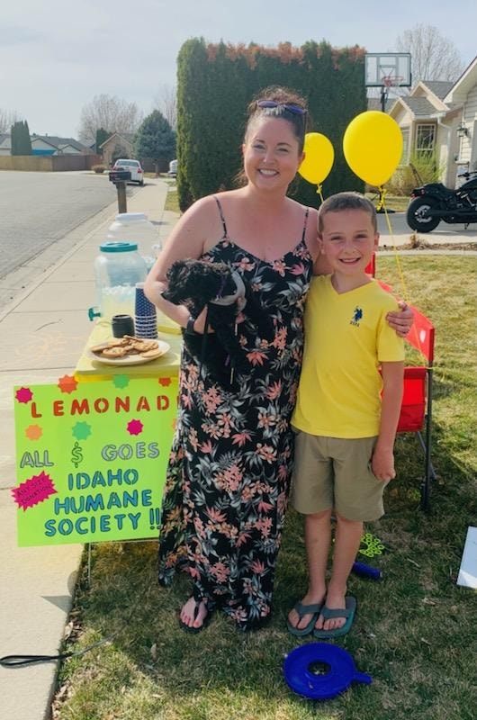 9-year-old ben miller smiling as he poses with a woman who is holding a small black dog. they are standing in front of ben's lemonade stand near the sign indicating that all sales go to idaho humane society.