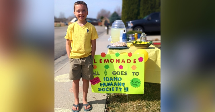 9-year-old ben miller smiling as he poses next to his lemonade stand in his front yard. directly next to him is a sign in the ground. it’s yellow and reads “lemonade. all $ goes to Idaho humane society !!!”