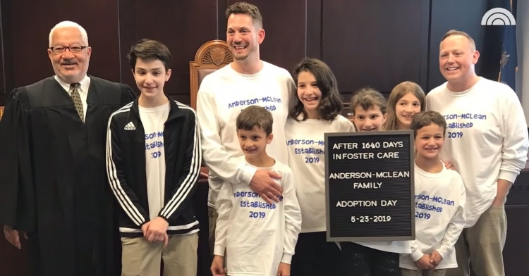 steve and rob anderson-mclean smiling as they pose with a judge and their newly adopted children: carlos, guadalupe, maria, selena, nasa, and max. they're all wearing the same white shirt that reads "anderson-mclean. established 2019." one of the children is holding a sign that reads "after 1640 days in foster care anderson-mclean family adoption day. 5-23-2019."
