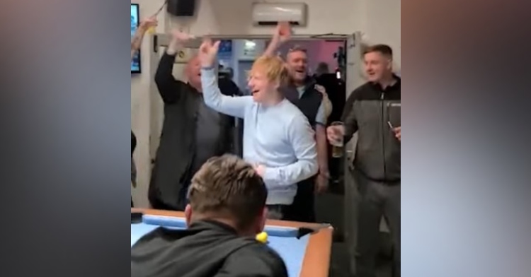ed sheeran smiling as he sings along with other patrons in a pub in small heath called the roost. he and several of the patrons around him have a hand raised in the air. they are standing near a pool table.