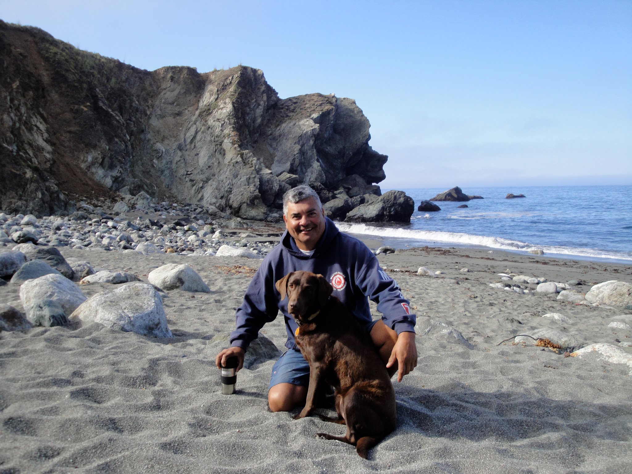 boat captain and fishermen michael arujo. he is on his knees, smiling, as he poses with his large, brown dog on a beach.