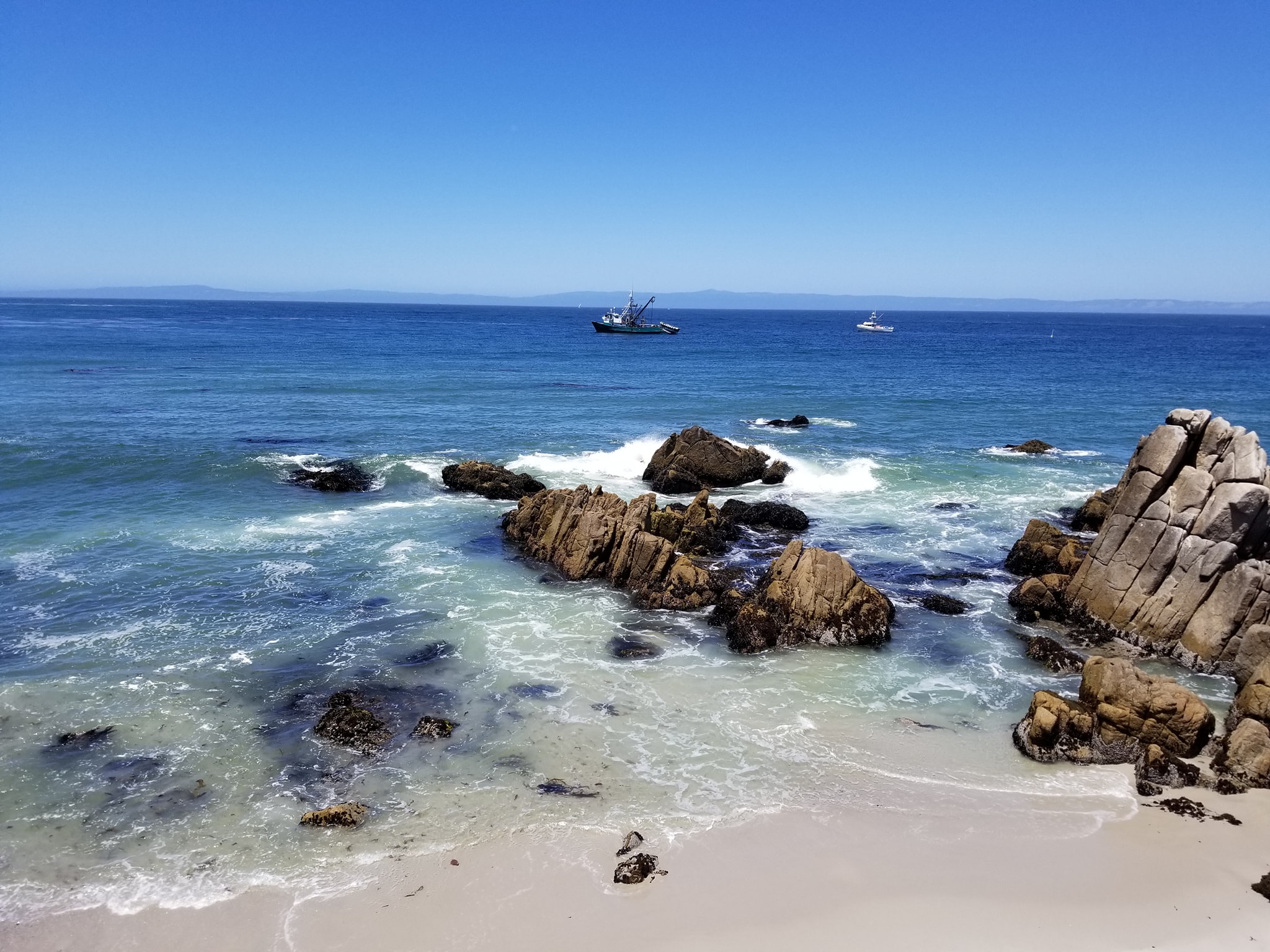 view of the monterey bay. there are rocks where the sea meets the shore and two boats on the water in the distance.