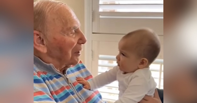 closeup of a 94-year-old man with alzheimer’s talking to his 6-month-old granddaughter, elsa, who is happily being held on his lap.