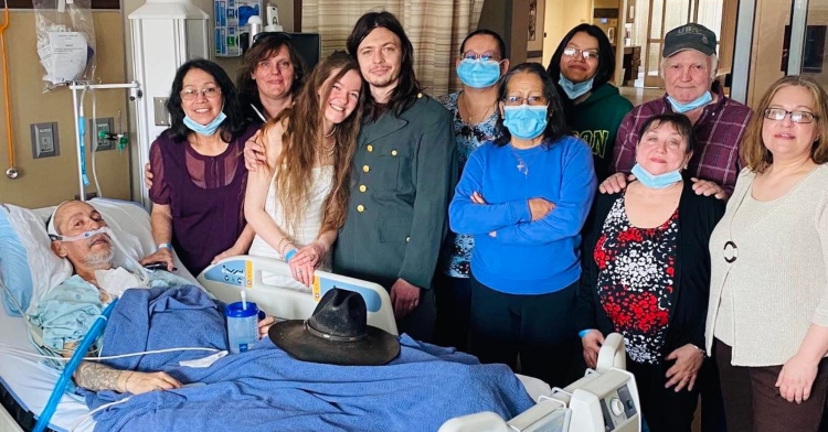 ethan and hailey carvey posing for a photo with friends and family after getting married in a hospital room. everyone is standing by jose guzman who is laying in a hospital bed.