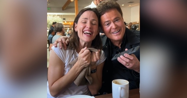 jennifer garner and donny osmond sitting together at a restaurant. osmond has one arm around garner and with the other he’s holding his phone. they’re both smiling as they sing, and garner has her eyes closed.