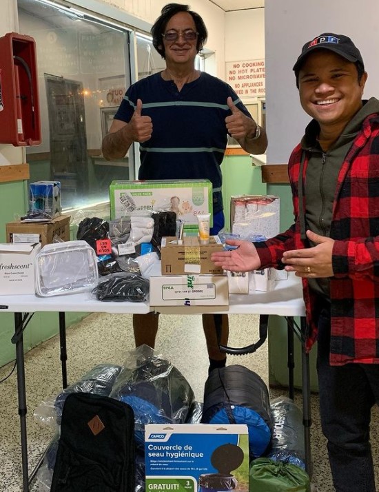 a man who goes by steven the maid and brian acosta arya smiling and posing together next to a table full of amazon donations for guests at their motel, lincoln tunnel motel