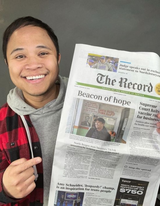 man named brian acosta arya smiling as he holds a newspaper with one hand and with the other points to the article about himself and his motel, lincoln tunnel motel, called "beacon of hope" 