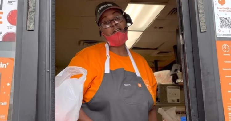 view of cynthia carter, a popeye’s cashier, as seen by a driver in their car. cynthia is standing at the drive thru window and is talking to the customer. she is wearing her popeye’s uniform and her headset.