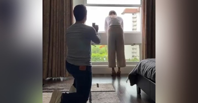 a man named chris lopez is on one knee as he holds out a box that contains an engagement ring. he is proposing to his girlfriend, susana, who is looking out of a window.