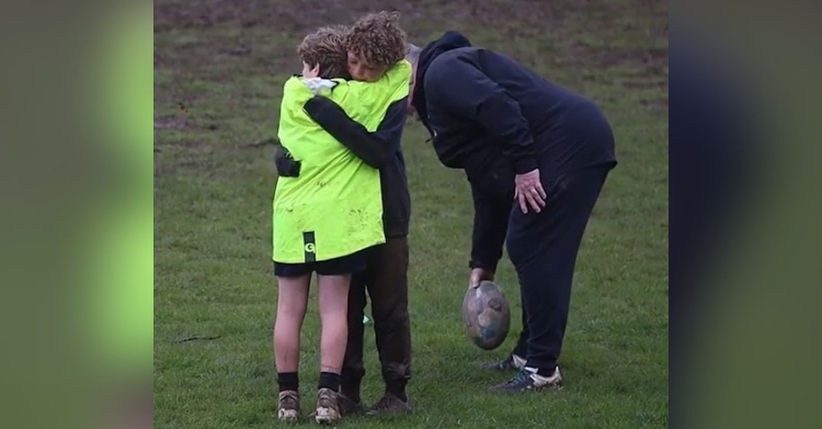 two young boys on the sedbergh school rugby team hugging as their coach grabs the ball off of the ground just behind them.