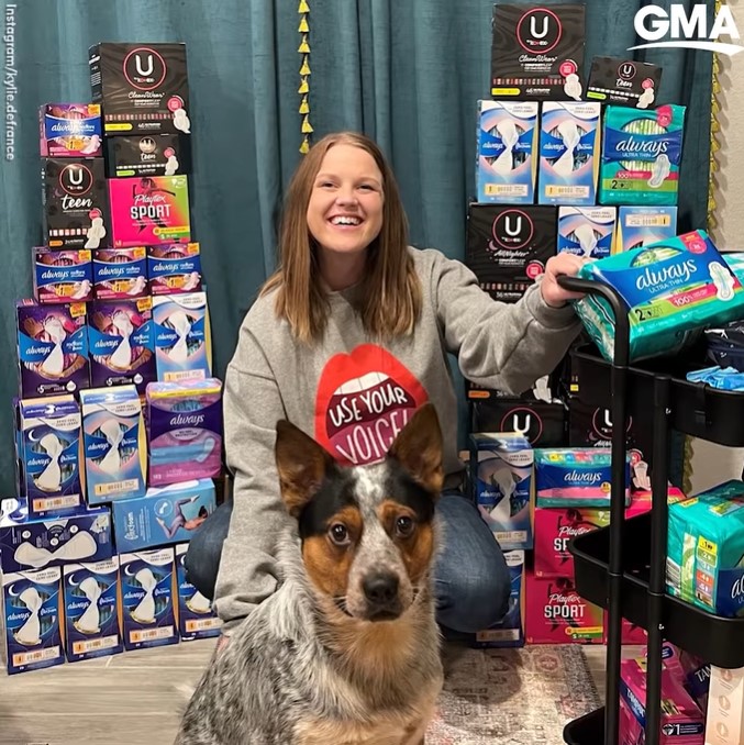 kylie defrance smiling as she poses with her dog. she is squatting as she holds on to a black cart next to her. the cart is filled with feminine hygiene products. there are feminine hygiene products neatly on display in stacks behind her, too.