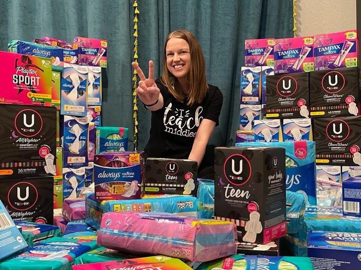 kylie defrance smiling and giving a peace sign as she poses at a table full of pads, tampons, and other feminine hygiene products that were donated to her classroom.