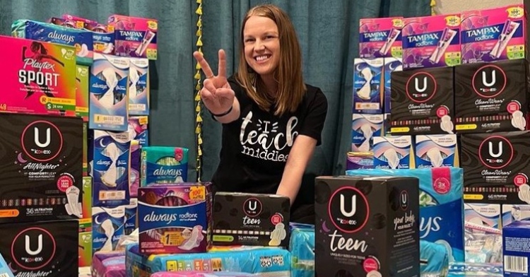 kylie defrance smiling and giving a peace sign as she poses at a table full of pads, tampons, and other feminine hygiene products that were donated to her classroom.