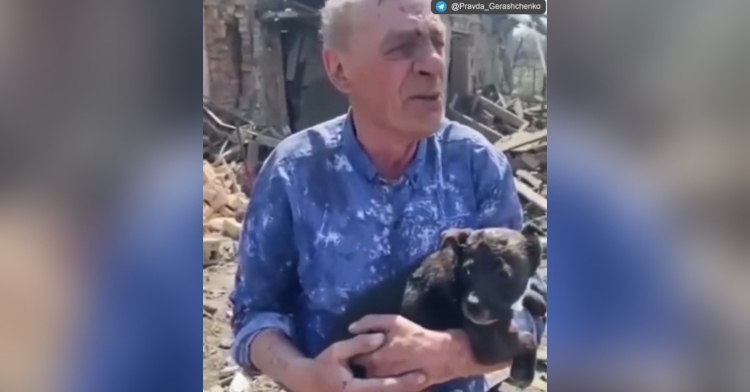 a ukrainian man holding his small, dark brown dog in his arms. he is standing amidst rubble in a village on dombas after a russian shelling.