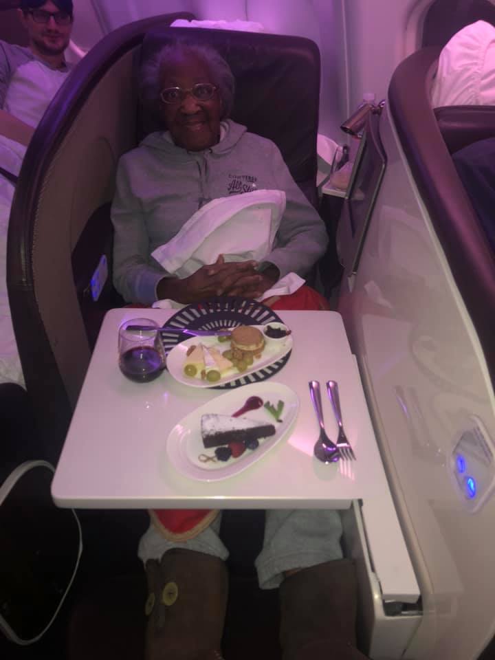 88-year-old violet enjoying first class from her seat. she's smiling. the table in front of her has plates of food.