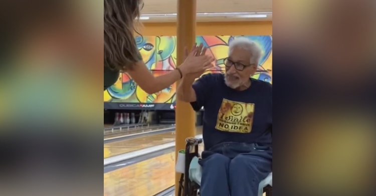 a woman high-fiving 90-year-old abuelito who smiles as he sits in his wheelchair after bowling.