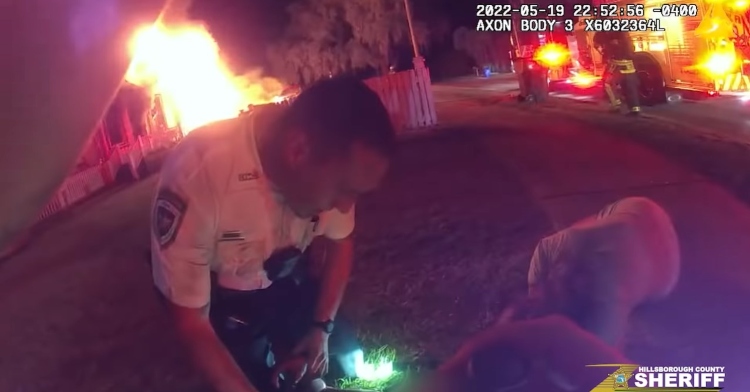bodycam footage of an officer looking down at a 9-year-old boy they rescued from a house fire. the flames can be seen in the distance behind them.