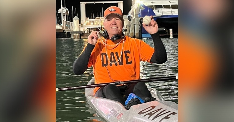 dave smiling as he sits in a kayak labeled “dave” while wearing an orange shirt that says “dave.” he’s holding up the pearl necklace around his beck with one hand and is holding the giants’ baseball he caught with the other.