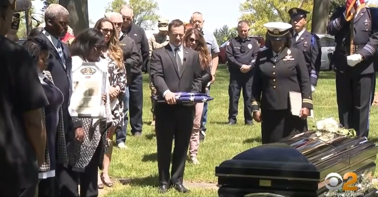 people gathered around eugene dedman’s casket at his funeral. one man is holding a folded up american flag while others stand nearby and hold flags.