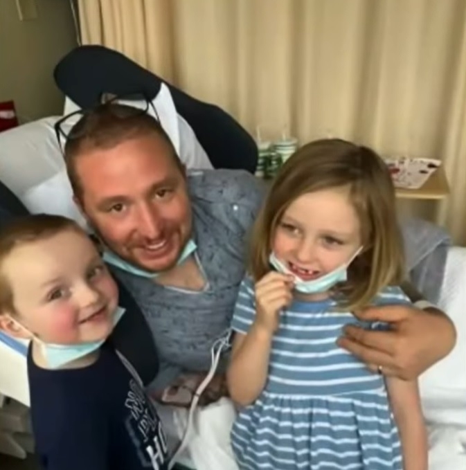 kyle semrau smiling as he lays in a hospital bed and poses with his kids. 