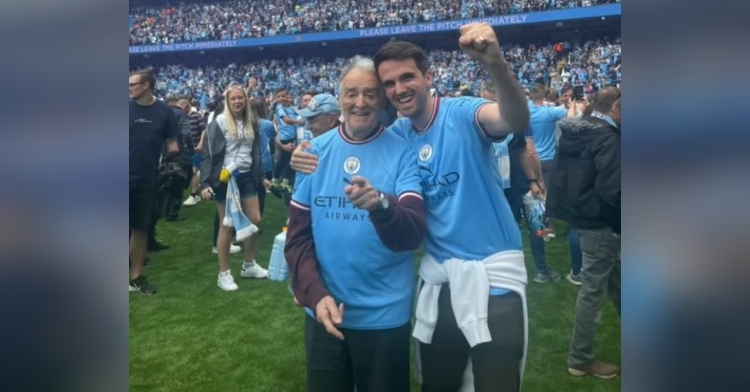 charlie gibson and his grandad smiling as they stand on a football field after a manchester city game. fans are standing near them in the background.