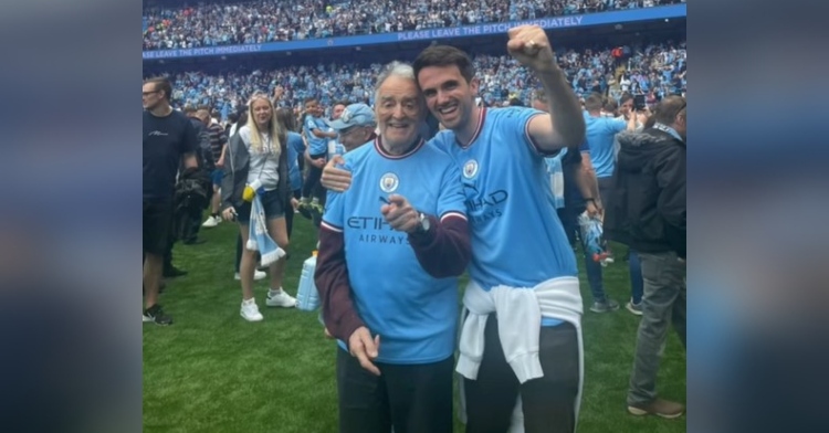 charlie gibson and his grandad smiling as they stand on a football field after a manchester city game. fans are standing near them in the background.