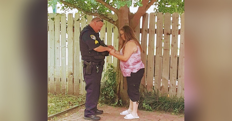 misti doyle and police officer sgt. truan holding hands and closing their eyes as they pray. they're standing outside next to a fence and underneath a tree.