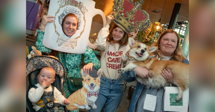 three women posing for a photo at the corgi café party. two are posing with large queen elizabeth themed props. one is holding a corgi. a baby sits in front of them in a stroller.