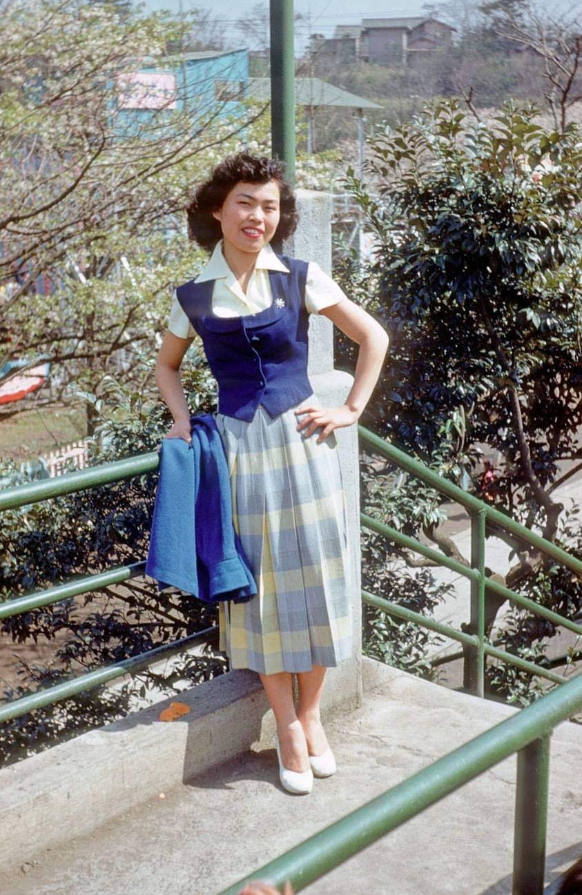 peggy yamaguchi smiling as she leans against a staircase railing outside next to trees. 
