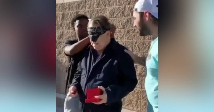 a man called mr. joe holding a key to his new truck as he stands outside, blindfolded. two high school students stand next to him. one has his hand on mr. joe’s shoulder and the other is untying his blindfold.