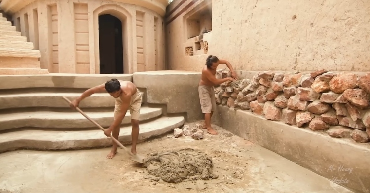 two men building an underground hut. one is shoveling cement-like material and the other is placing rocks on an outside wall.