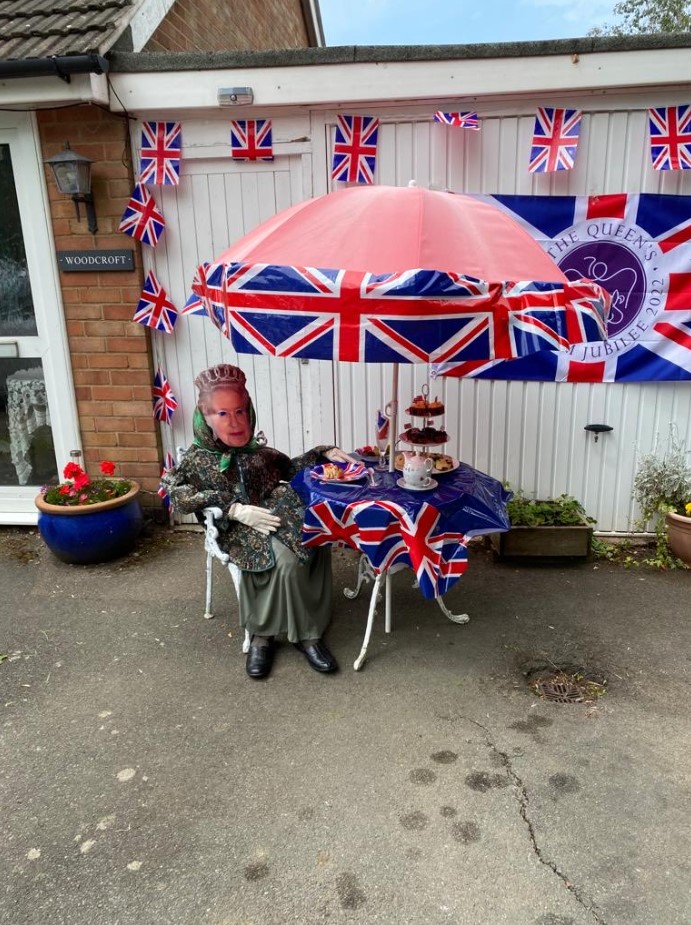 scarecrow with queen elizabeth's face that's sitting at a small table with an umbrella outside. the umbrella, table, and outside wall are decorated with the union jack. there's pastries and teacups on the table.