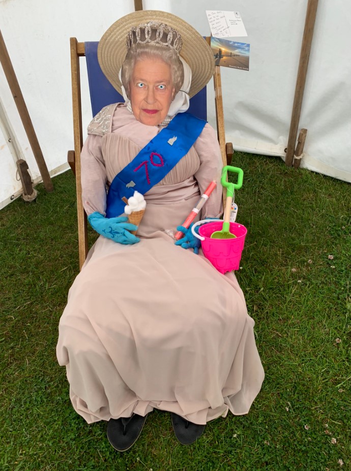 scarecrow with queen elizabeth's face sitting on a lawn chair. she has a crown, sash, fake ice cream cone, and a pail and shovel.