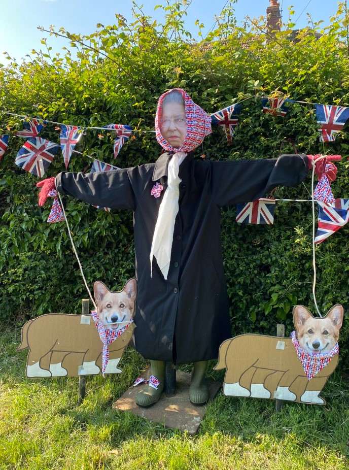 scarecrow with queen elizabeth's face stood in front of greenery that's decorated with the union jack. two cardboard corgis with photos of real corgi faces glued on them are on either side of the scarecrow.