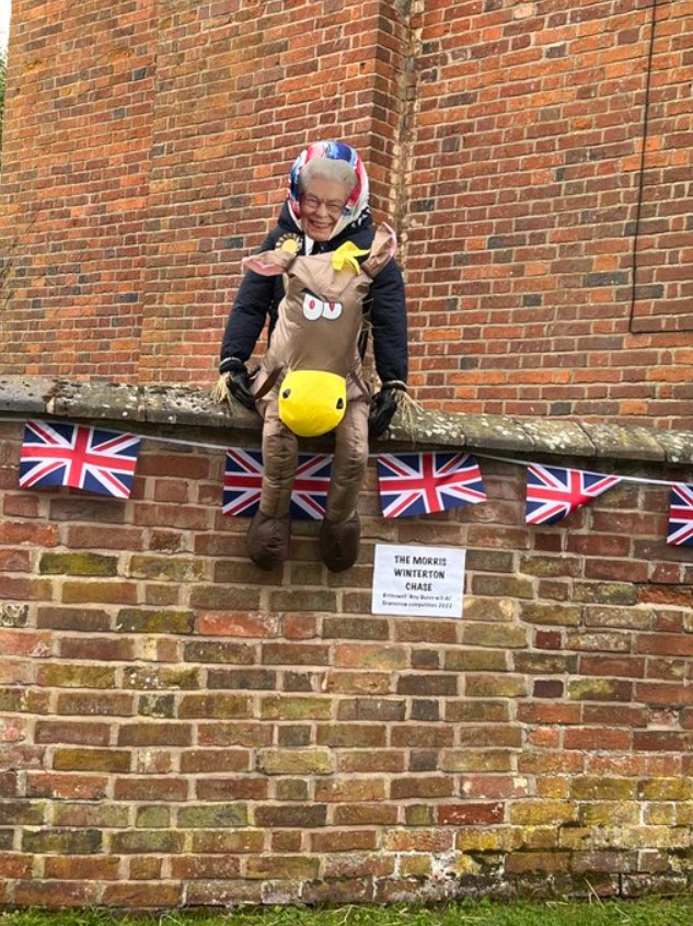 scarecrow of queen elizabeth sat on a brick wall with british flags hanging on it. the scarecrow has queen elizabeth's face and she's on a donkey.