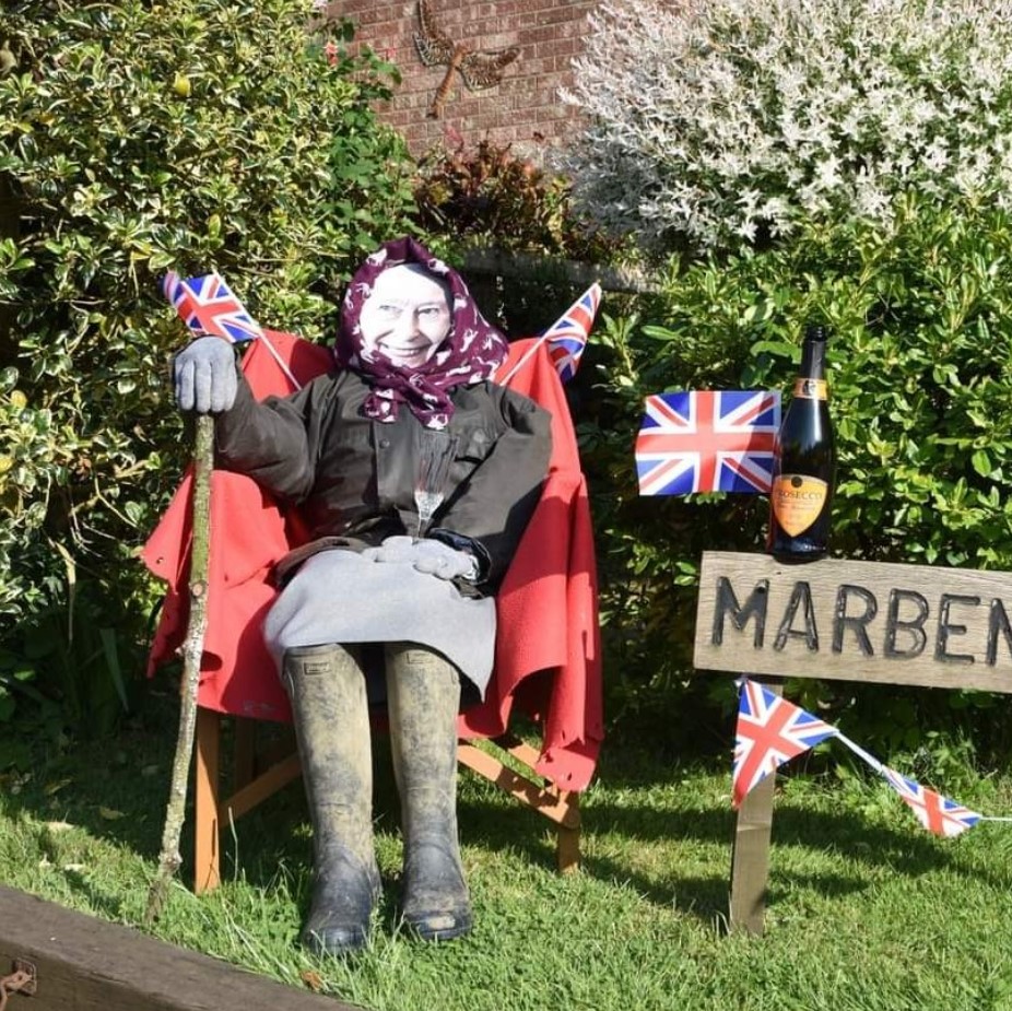 scarecrow with queen elizabeth's face sitting outside in a chair next to british flags.