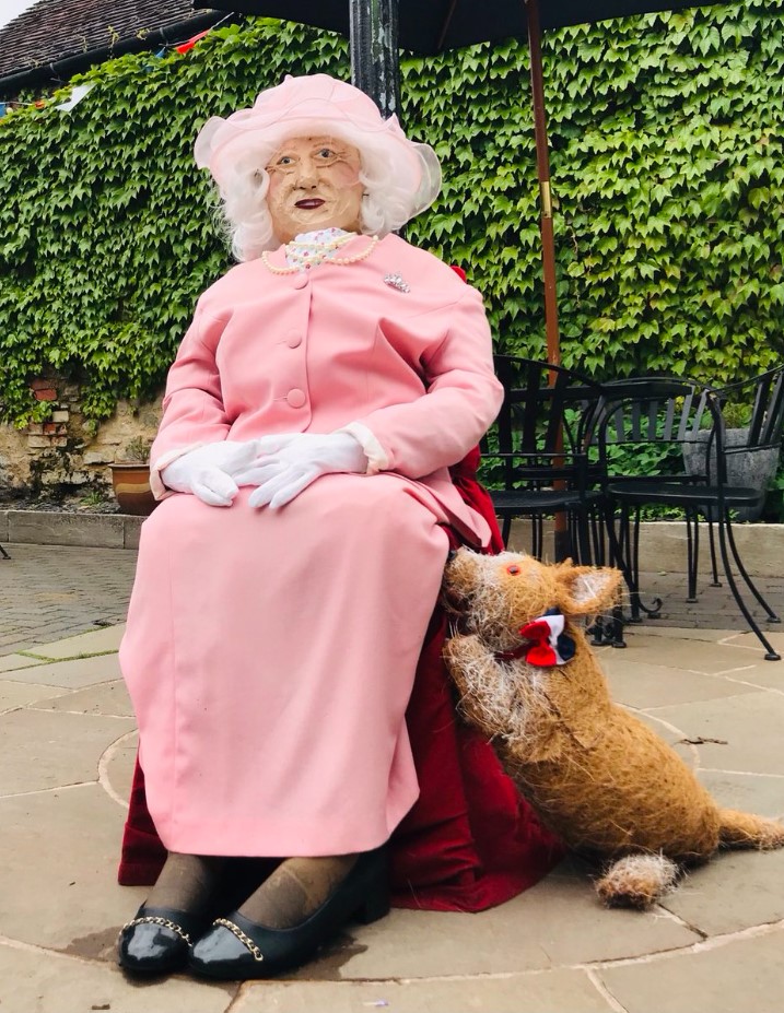 scarecrow of queen elizabeth that is sitting outside. the scarecrow is dressed in a pink outfit, gloves, and has scarecrow a corgi next to her.
