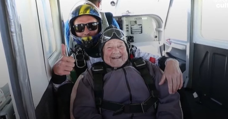 rut larrson and her parachute buddy posing for a photo in the plane they’ll be parachute jumping from. he’s wearing sunglasses and is giving a thumbs up and she’s smiling.