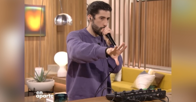 mohamed belkhir performing for canal+ using a loop station. he’s standing at a table and is using a microphone that he’s holding with one hand. with he other he’s reaching out into the air directly in front of him. behind him is a living room with a brown and yellow color scheme.