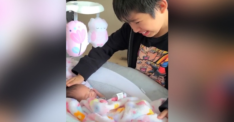 little boy crying as he gently touches his baby sister’s head. the baby is laying in a basinet.