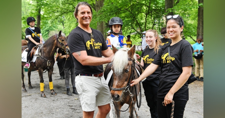 three people wearing gallopmyc shirts smiling as they stand around a horse with a small child on top. behind them is another person on a horse with others nearby.